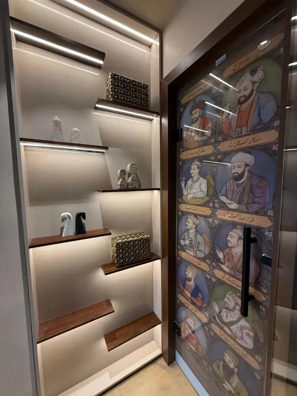 Back-lit shelves of brassware beside a block-printed panel. Tajnuma, Agra.